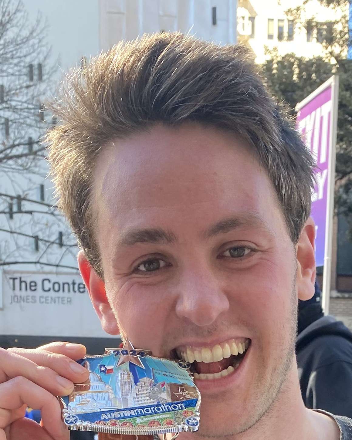 Richard Joseph Spinelli holding his 2024 Austin Marathon medal in his teeth, grinning, with city buildings behind him